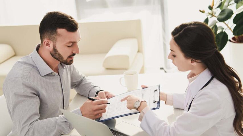 man signing magellan health insurance claim form for addiction rehab while doctor pointing at it in office with laptop
